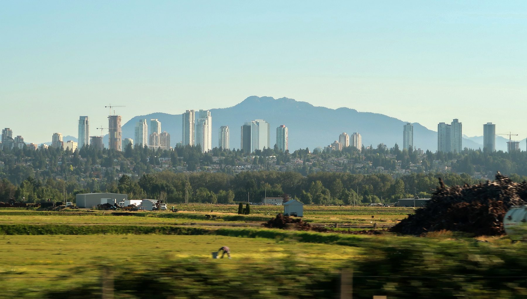 Metrotown_201807 City skyscrapers backed by mountains, overlooking foreground farmland and forest.